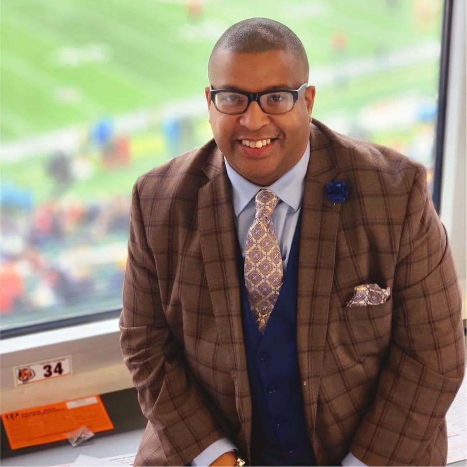 Darren Smith, a Kansas City sports broadcaster, is smiling while seated in a press box overlooking a sports field. He is wearing a brown plaid suit with a blue vest, a patterned tie, and glasses.