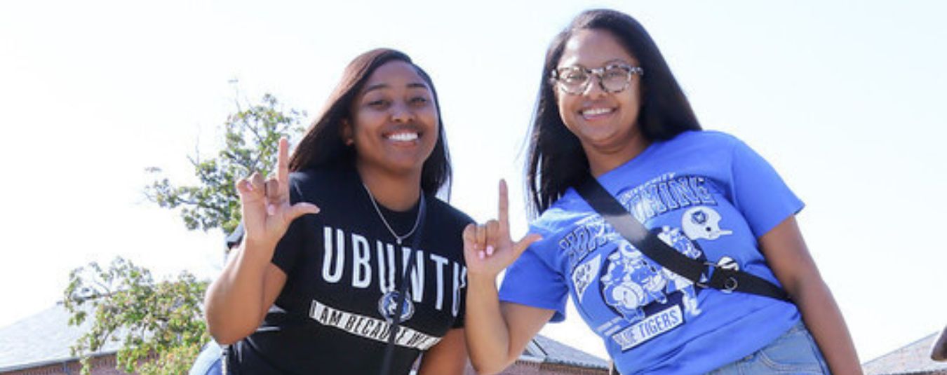 Two young women smiling outdoors, making hand signs. One wears a 'UBUNTU' shirt, the other a 'Lincoln University Blue Tigers' shirt.