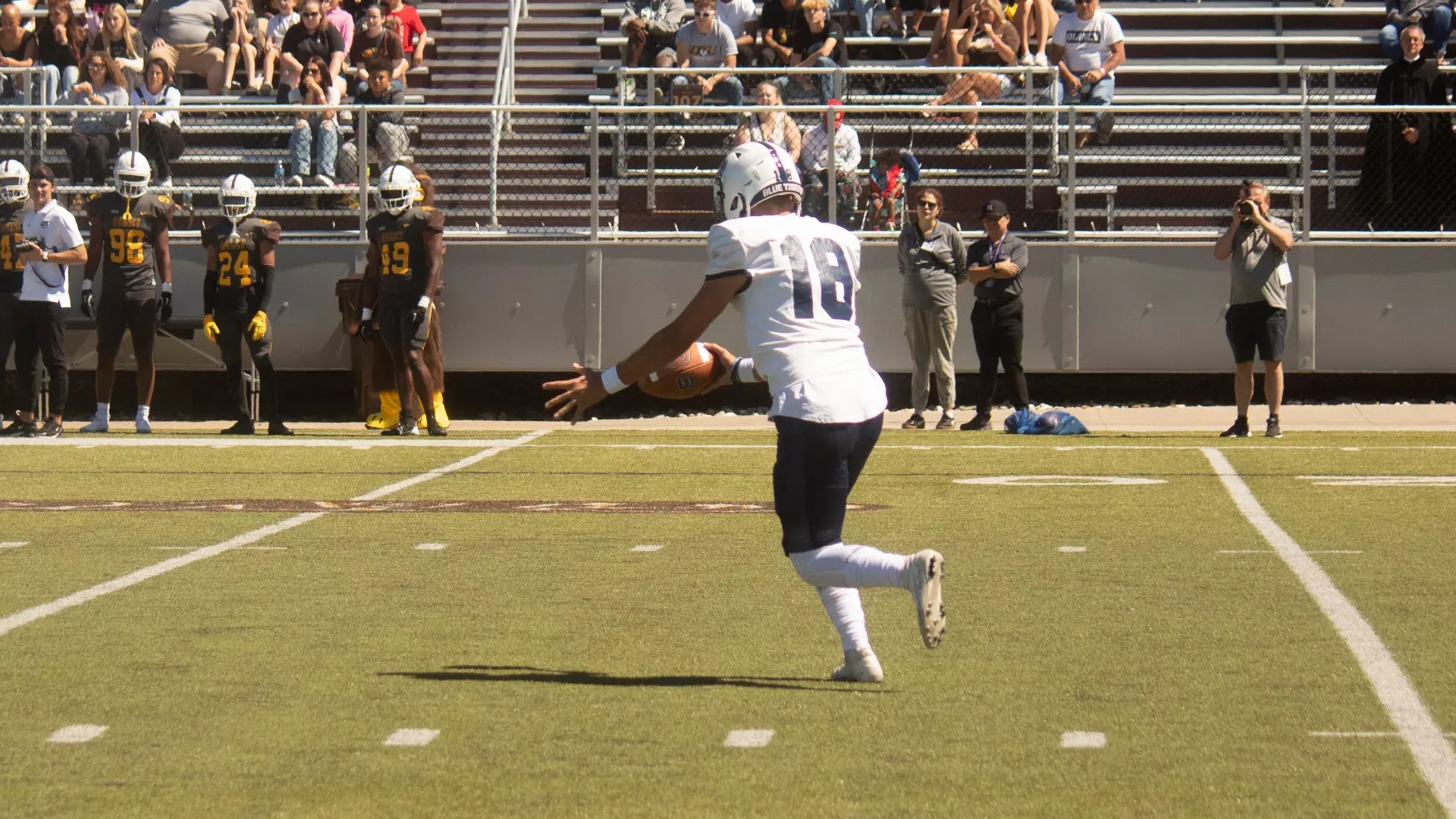 Clayton Winkler, in a white Lincoln University football jersey (#18), is holding the football for a field goal attempt while a teammate prepares to kick during a game, with opponents and officials visible in the background.