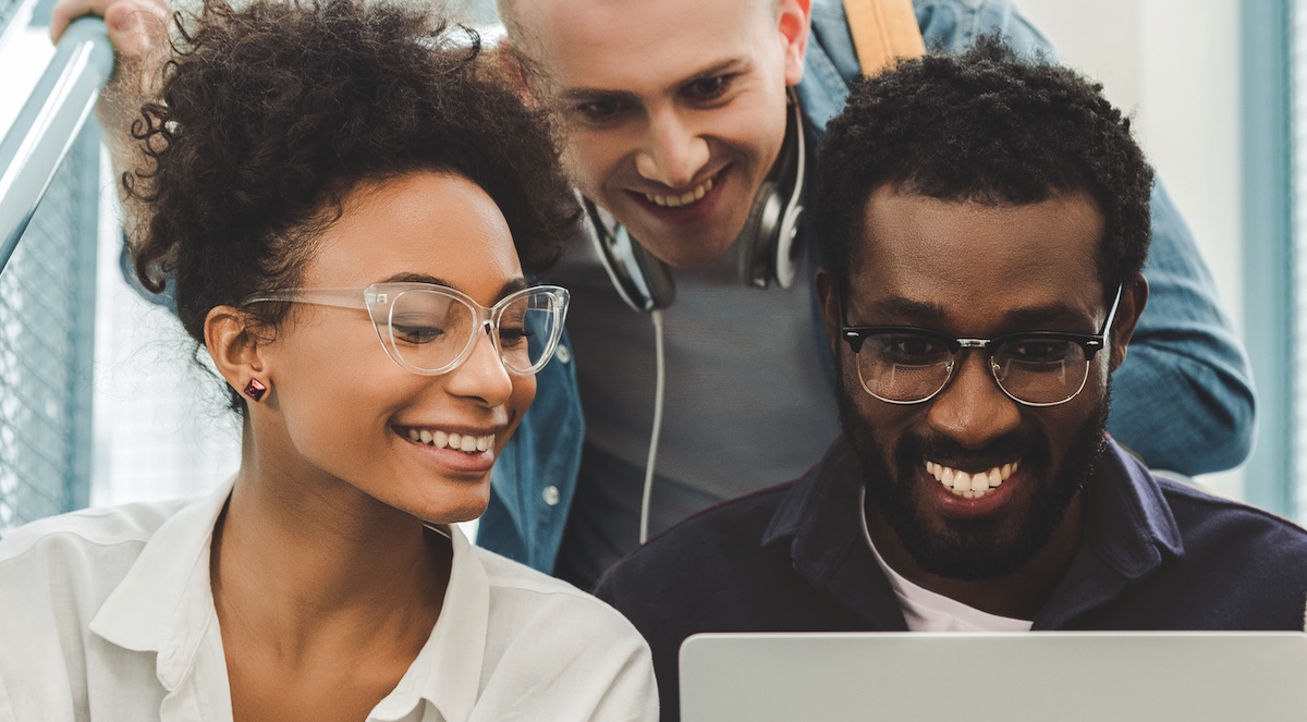 Three students—one female and two male—smile while looking at a computer. 