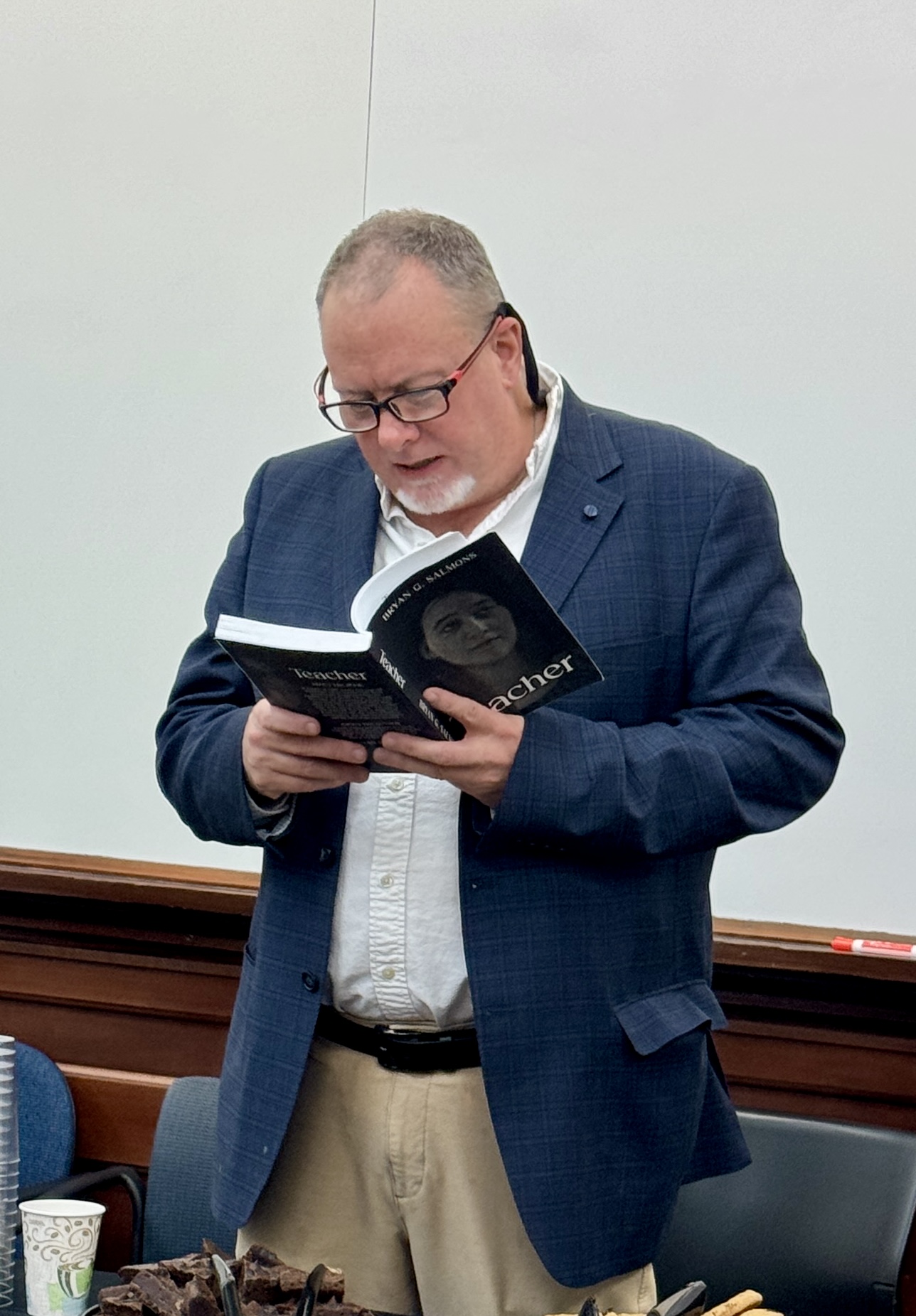 A middle-aged man with short, graying hair and glasses is reading aloud from a book titled Teacher in a classroom setting. He is wearing a navy-blue blazer over a white dress shirt. A table in front of him has cups, plates, and snacks. The background consists of a whiteboard and a classroom wall.