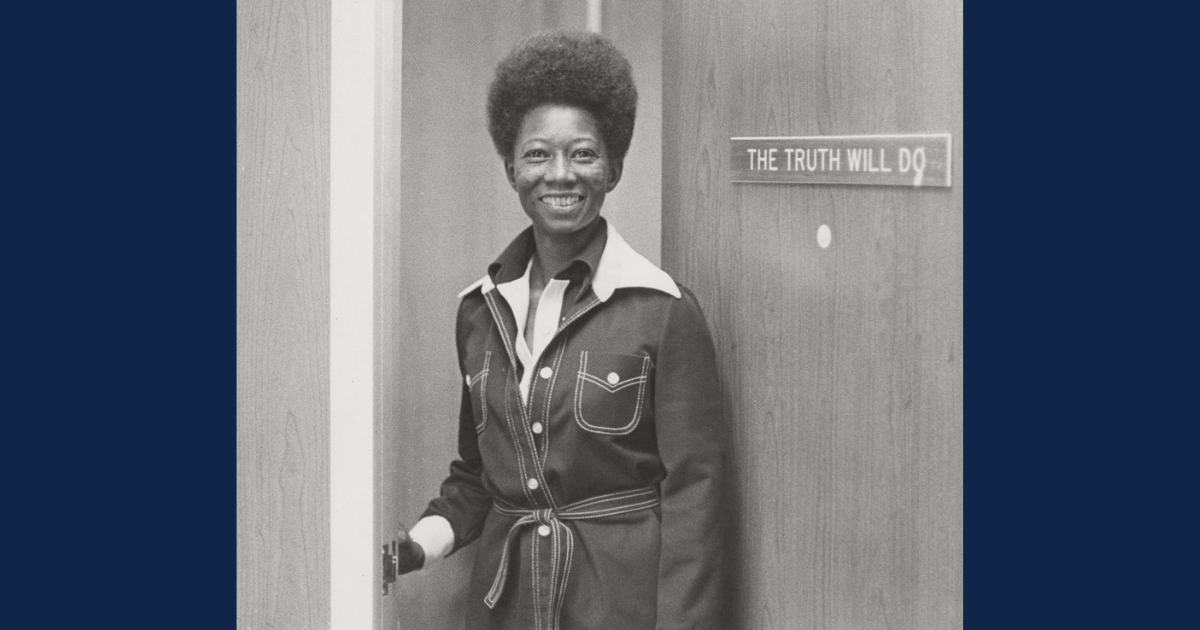 A black-and-white photograph features a smiling woman with short, natural hair standing in a doorway. She is wearing a dark-colored, belted jacket with contrast stitching and a wide collar. The sign on the door behind her reads, "THE TRUTH WILL DO." The background consists of a wooden door and a light-colored wall. The woman appears confident and warm, engaging directly with the camera.