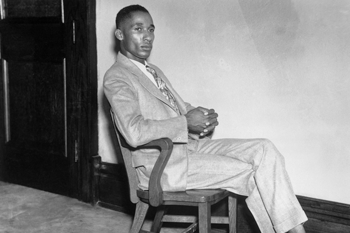 Black-and-white photograph of Lloyd Gaines, a young African American man, sitting in a wooden chair. He is dressed in a light-colored suit with a patterned tie, his hands clasped together, and his legs crossed. His expression is serious as he looks slightly off-camera. The background features a plain wall and a wooden door.