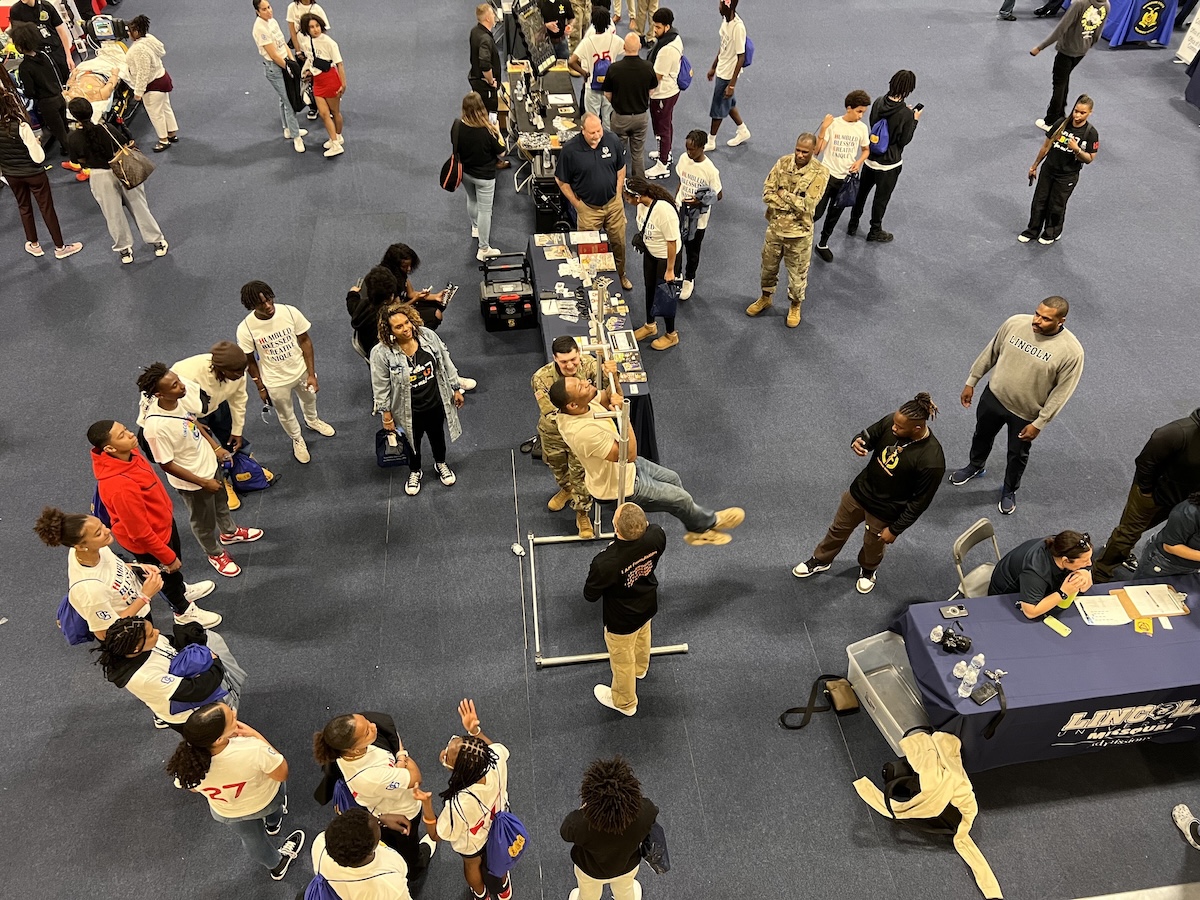 A large indoor career expo is underway with students and professionals engaging at various booths. Many attendees are wearing white t-shirts with blue text and carrying blue bags. A man in military fatigues stands near a table, while another person demonstrates a physical activity. Lincoln University branding is visible on a table in the foreground.