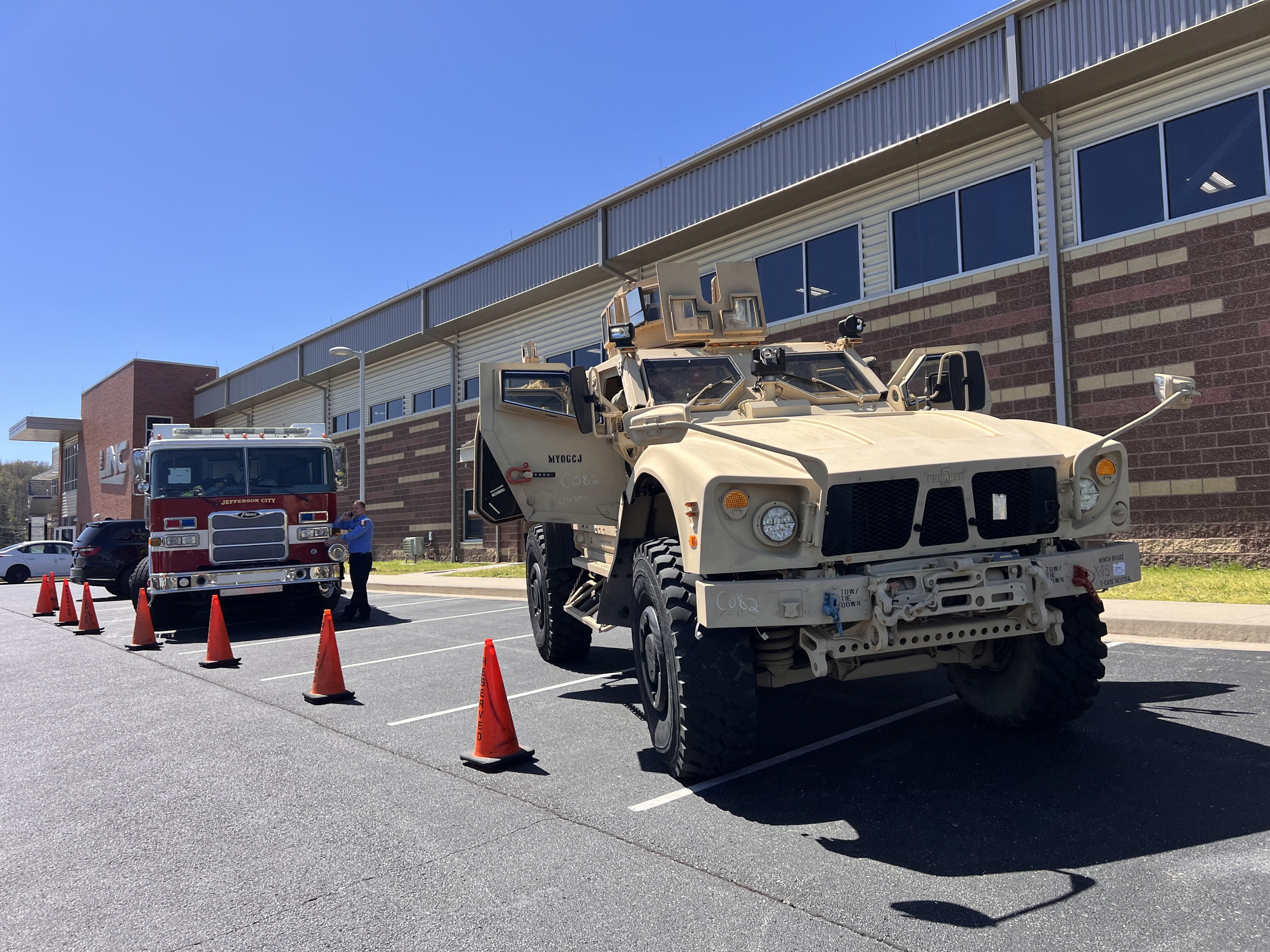 A tan military vehicle and a red fire truck are parked outside a brick building, with orange cones placed in front of them.