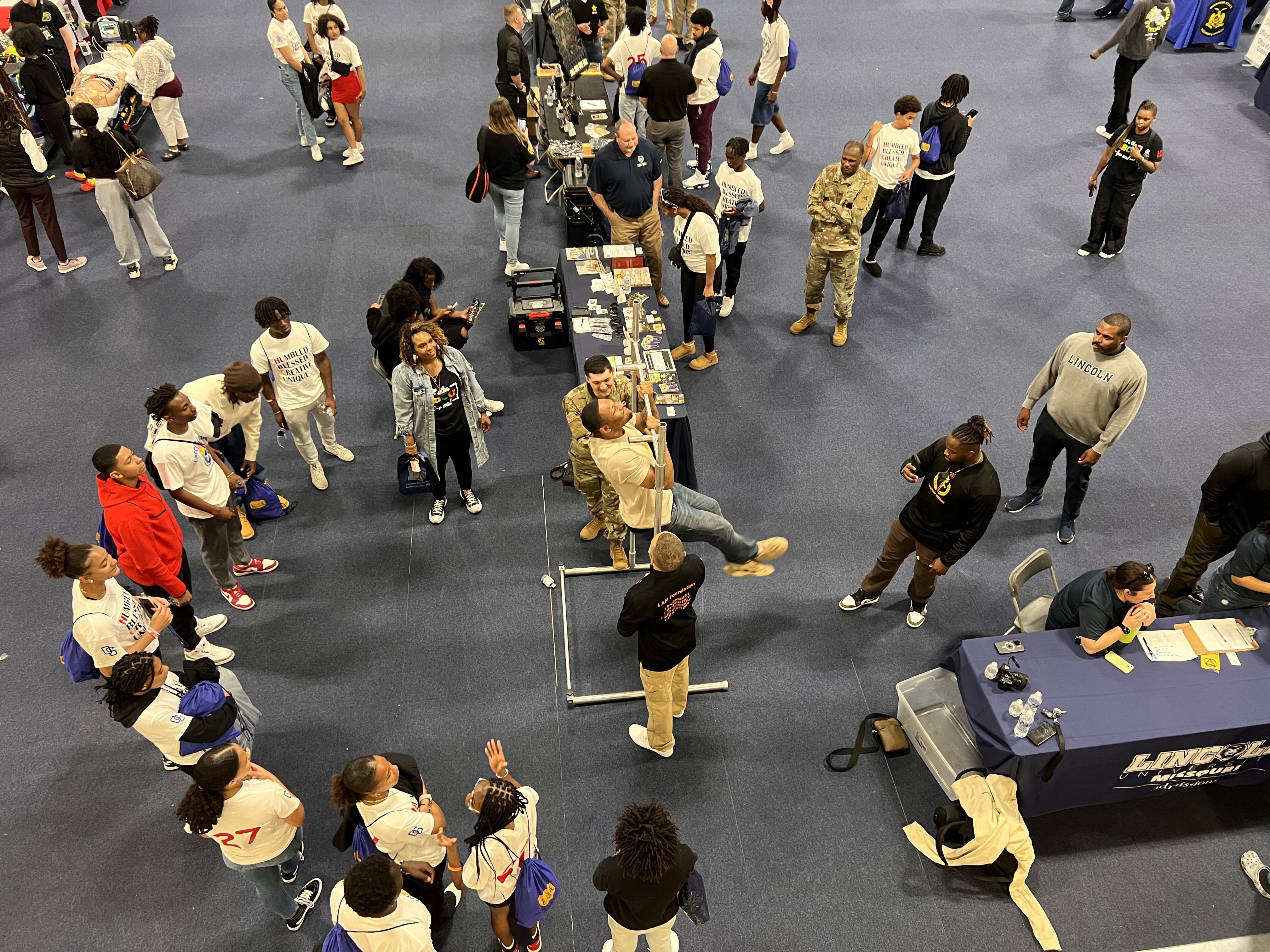 A career expo with students in white shirts, military personnel, and recruiters engaging at booths in a large indoor venue.