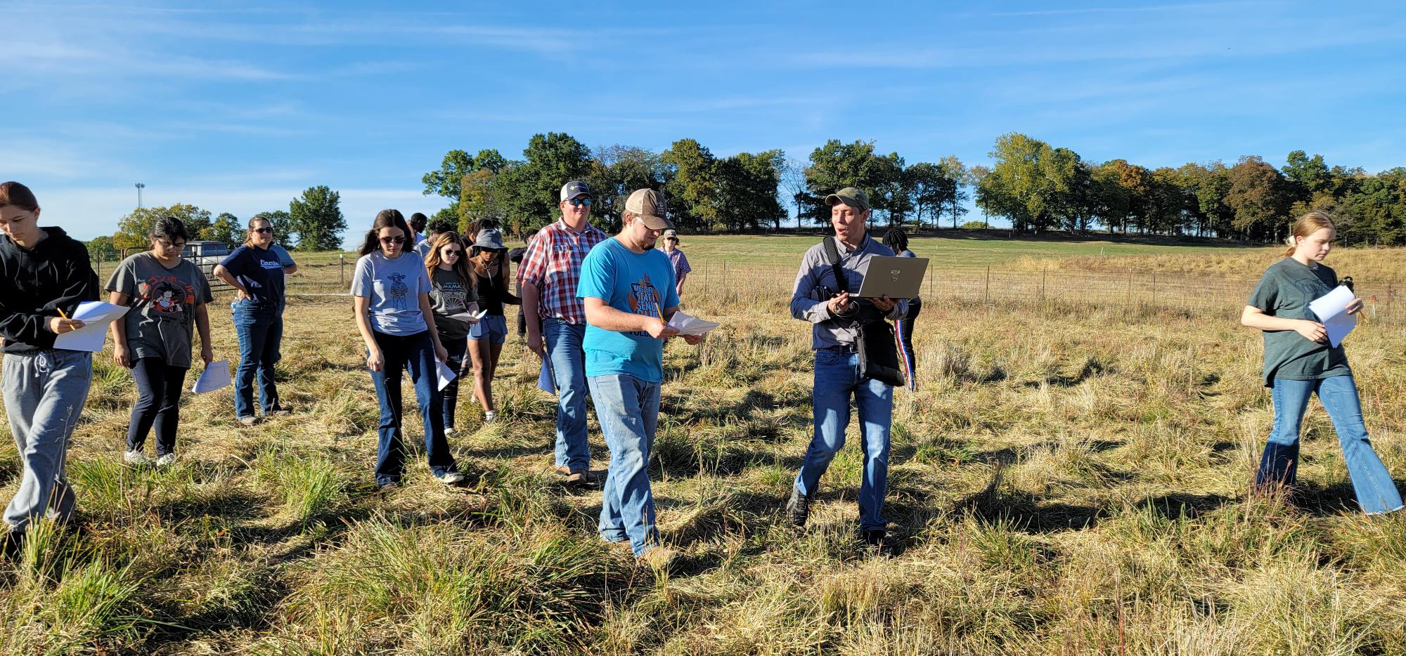 A group of students walks through a grassy field at Carver Farm at Lincoln University carrying clipboards and papers. Trees are in the background, and the sky is clear and blue.