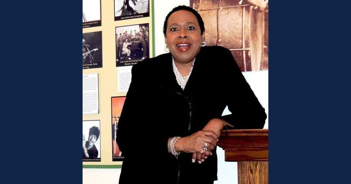 Florene Calvin Dawkins stands smiling at a podium, dressed in black, with a historic display featuring Louis Armstrong behind her.