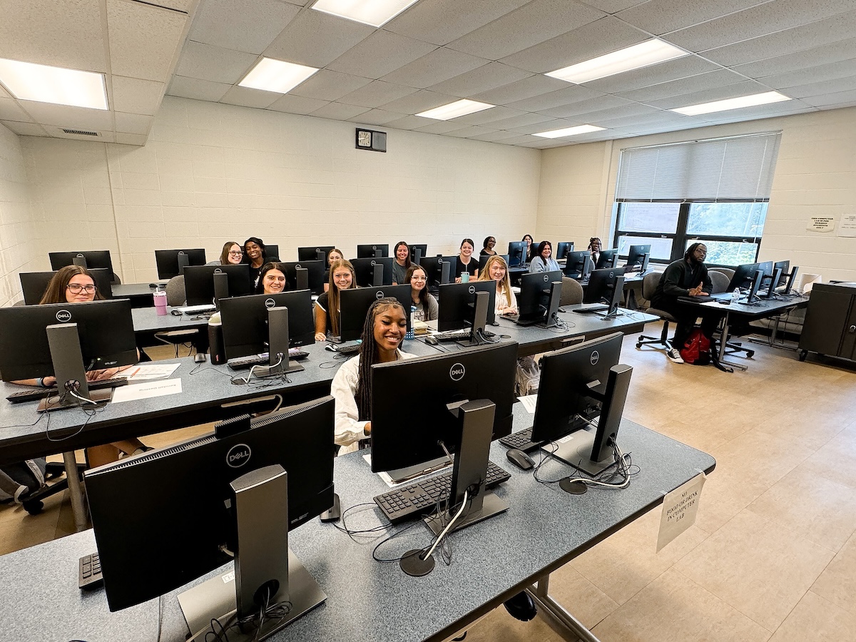 Students seated in a classroom filled with desktop computers, smiling and looking toward the camera. The room has a bright and welcoming atmosphere with large windows letting in natural light.