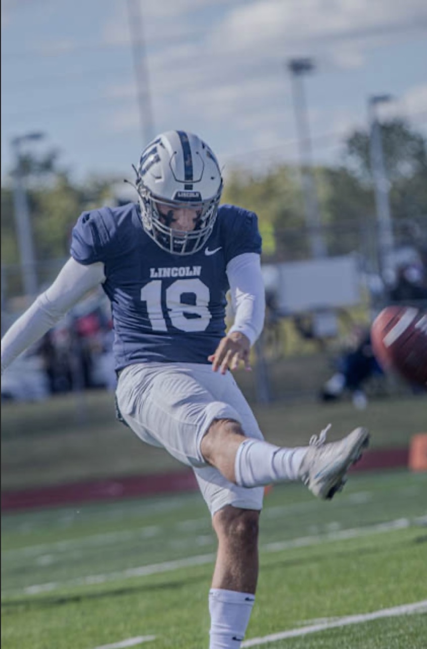 Clayton Winkler, a football player for Lincoln University of Missouri, wearing a blue jersey (#18) and white pants, is captured mid-action as he kicks a football during a game under a clear sky.