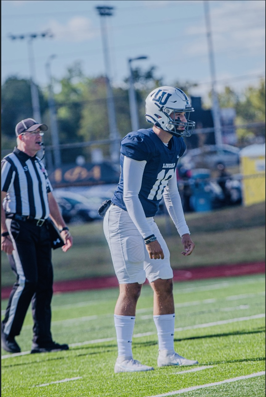 Clayton Winkler, a football player for Lincoln University of Missouri, wearing a blue jersey (#18) and white pants, is seen standing on the field during a football game with a referee behind him. 