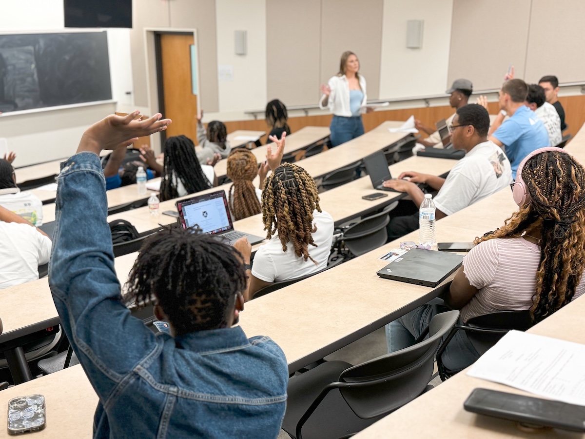 Students in a classroom actively engage with a lecture, with several raising their hands to participate. A female instructor stands at the front, addressing the class, while students use laptops and notebooks to take notes. The setting features tiered seating and a collaborative learning environment.