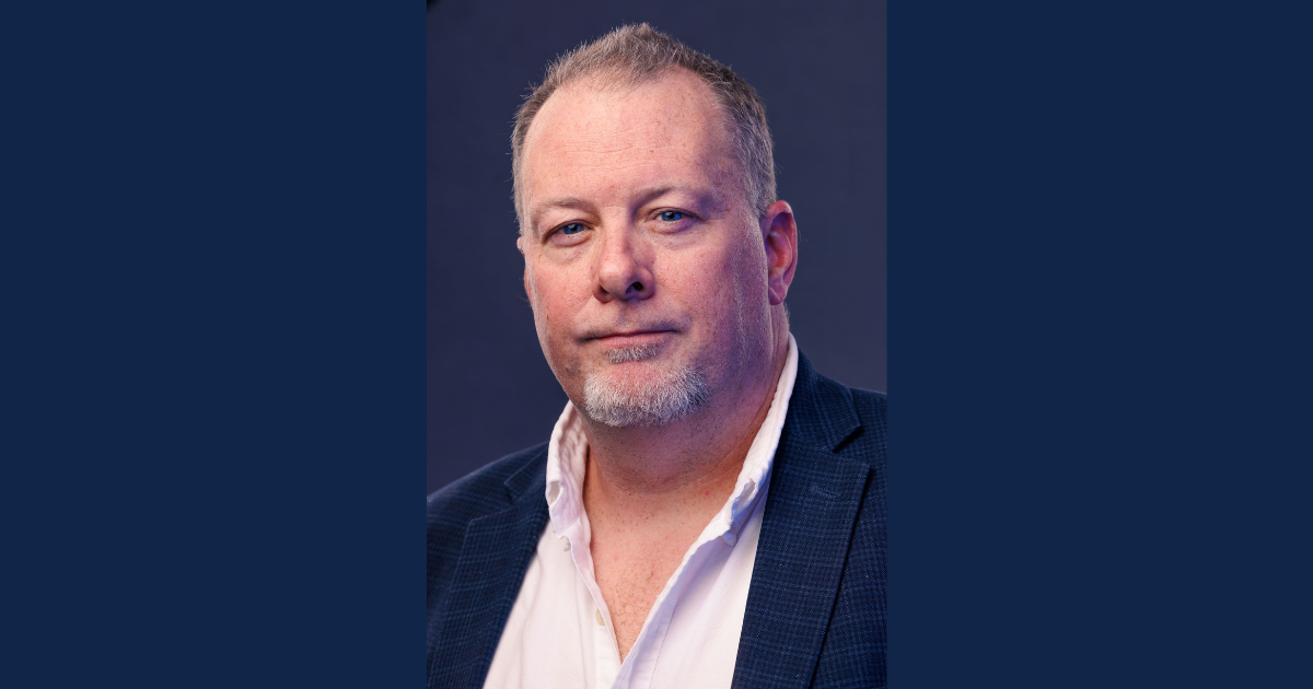 A middle-aged man with short, graying hair and a goatee wearing a navy blue blazer over a white dress shirt. He has a confident, relaxed expression with a slight smile, looking directly at the camera. The background is a solid dark blue.