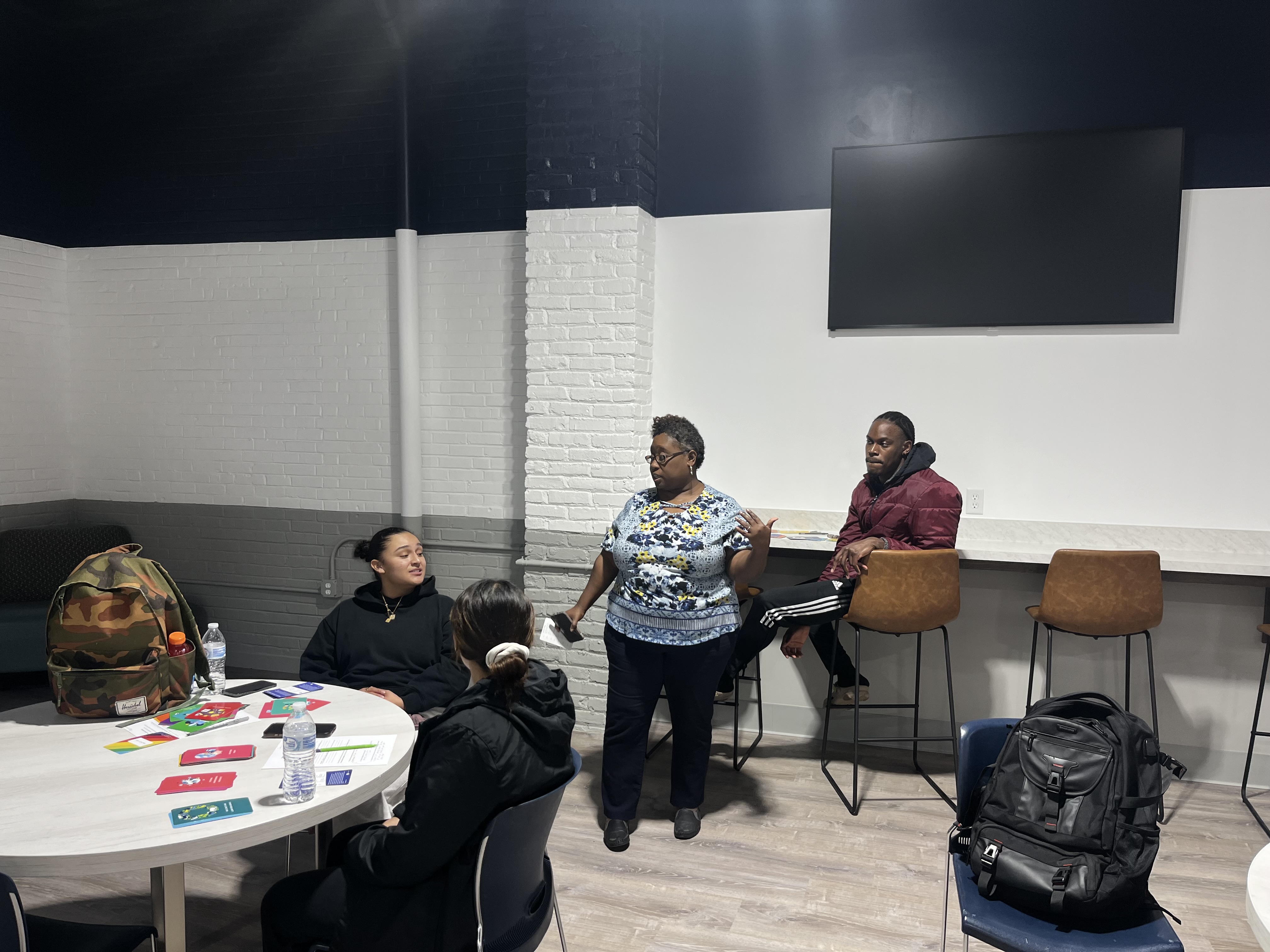  A woman in a floral shirt stands and speaks to two students seated at a round table in a modern lounge area. The table is covered with colorful cards and papers. A male student in a red jacket sits on a high chair against a white and navy-blue wall with a mounted TV. Backpacks and water bottles are visible on the tables.