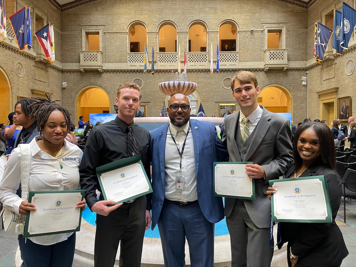  Four well-dressed student scholars stand with their USDA liaison displaying certificates of recognition.