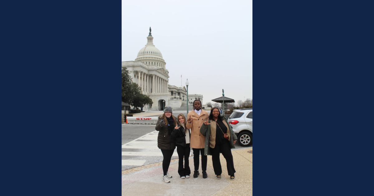 A group of four Lincoln University students stands in front of the U.S. Capitol building on a cloudy day. They are posing on a sidewalk, smiling and making the LU hand gesture. The students are dressed warmly, wearing coats and jackets. Cars and trees are visible in the background.