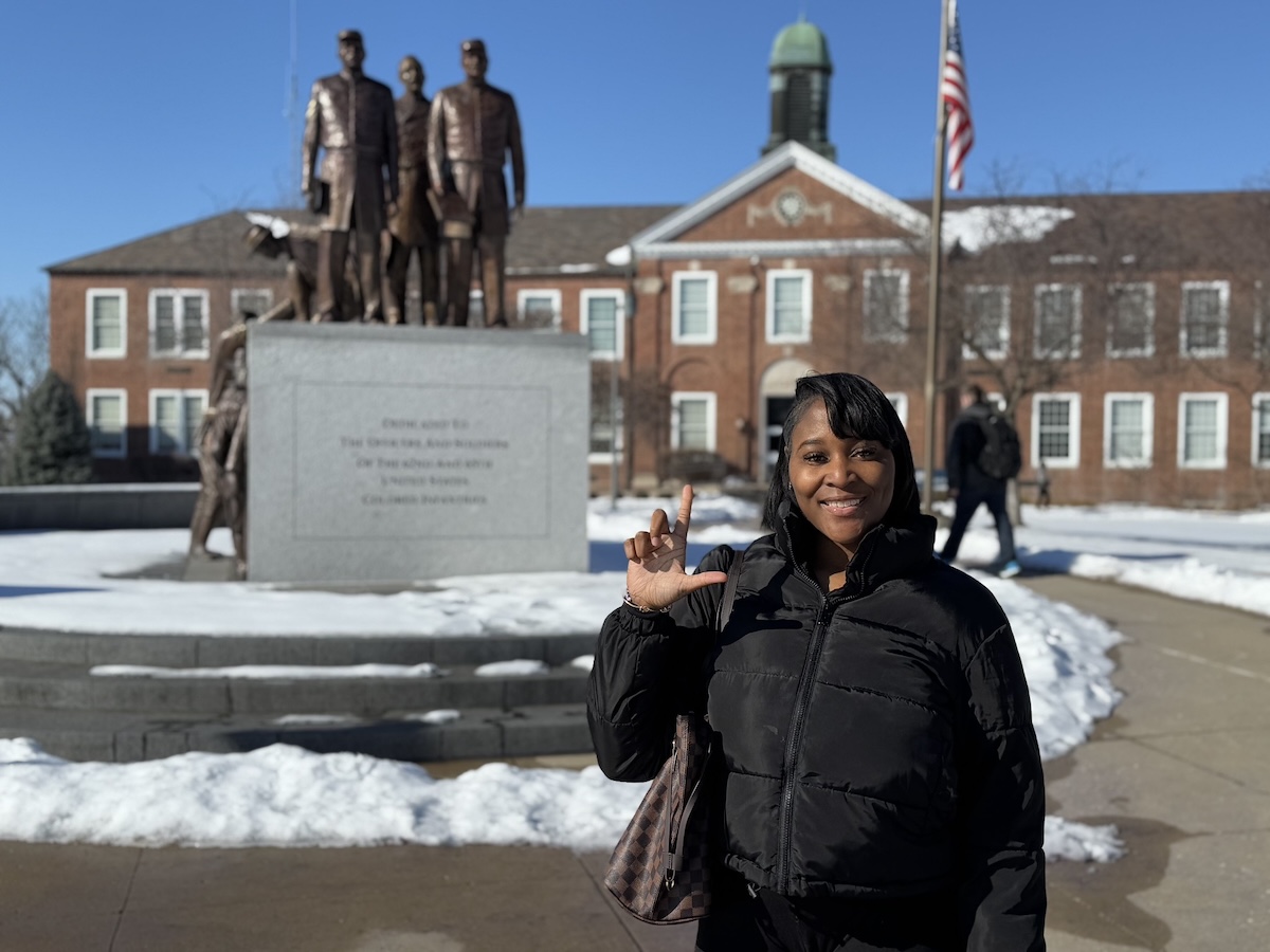 A female Lincoln University of Missouri student smiles warmly while standing in front of a statue and a brick building on a snowy campus. She is wearing a black puffer jacket and holding a handbag, making a hand sign with her right hand. The building in the background features a green-domed cupola and an American flag, with a clear blue sky above.