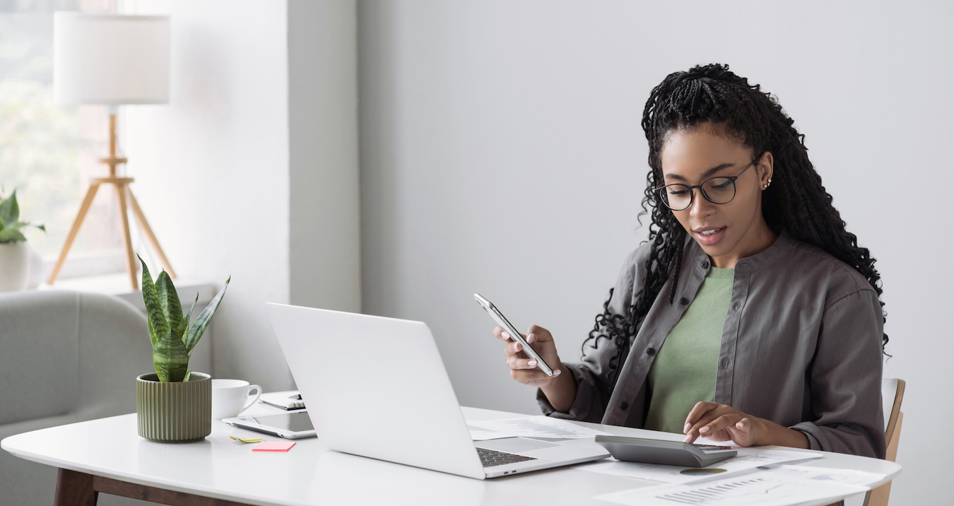 Young woman with glasses working at a desk, holding a phone, using a calculator, and reviewing documents next to a laptop and plant.