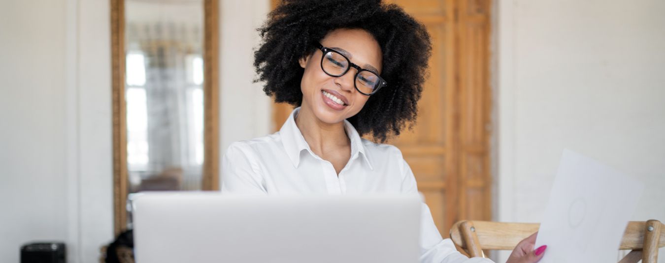 Smiling woman with glasses working at a laptop, holding a document, with a tablet and notebook on the table in a bright room.