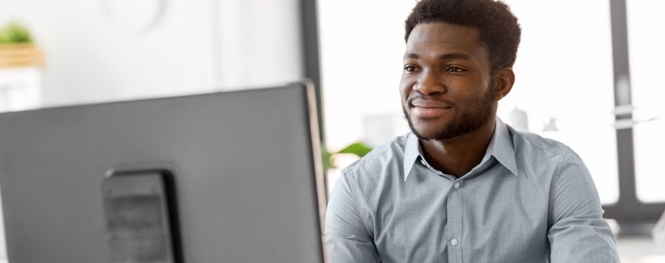 Man in a light gray shirt working at a desktop computer, smiling and typing, with office supplies on the desk in a bright workspace.